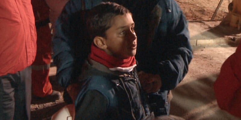 A young boy with a red scarf around his neck stands surrounded by people wearing blue and red jackets.