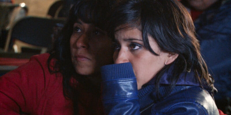 Two women with dark hair sit side-by-side, looking intently toward the right, with one woman wearing a red jacket and the other a blue leather jacket.