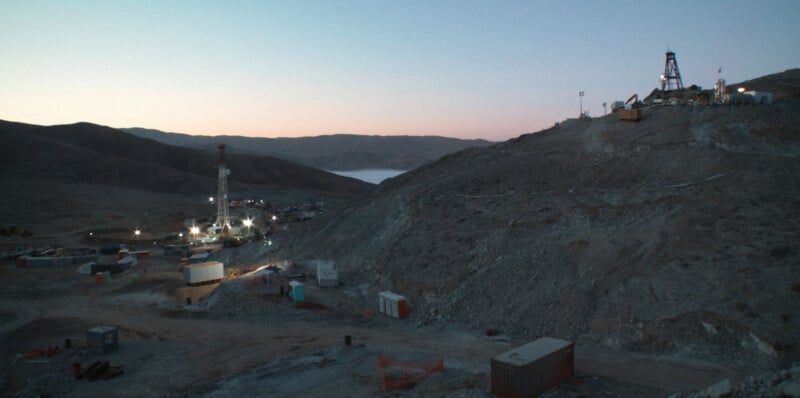 A drilling rig and industrial equipment sit in a desert valley at dusk, with a smaller structure on a hill to the right.