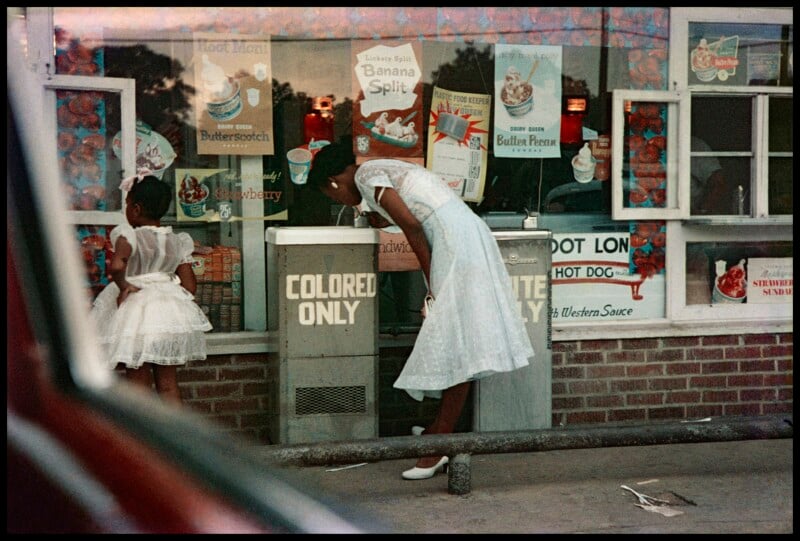 A woman in a white dress drinks from a water fountain labeled "Colored Only" while a young girl in a white dress stands nearby.