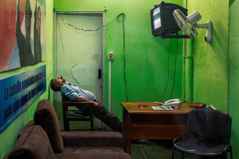A man leans back in a chair with his head tilted upward in a room with bright green walls, a wooden desk, and a wall-mounted television.