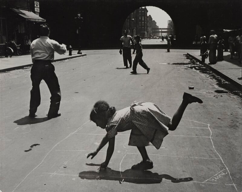 A young girl in a checkered dress leans forward to retrieve a marker from a hopscotch grid drawn on a city street, while a man stands nearby and others walk in the background under a large stone archway.