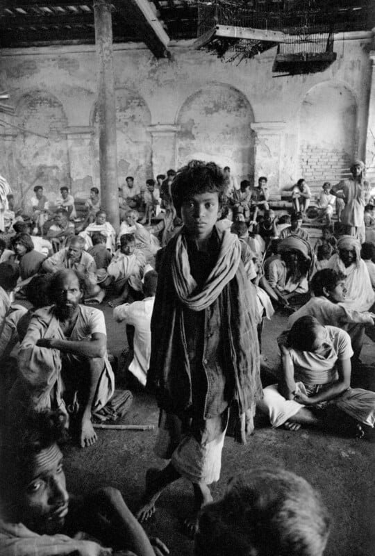 A young boy stands in the center of a crowded, dimly lit room filled with seated men, with birdcages hanging from the ceiling.