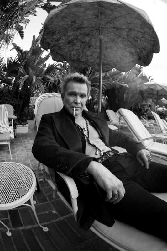 Billy Idol sits in a lounge chair under a patio umbrella, wearing an open black jacket, dark pants, and jewelry, with a cigarette in his mouth.