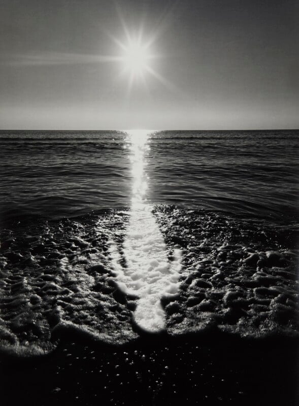 A bright sun shines above the horizon, casting a vertical reflection across the dark ocean water onto the foamy shoreline.