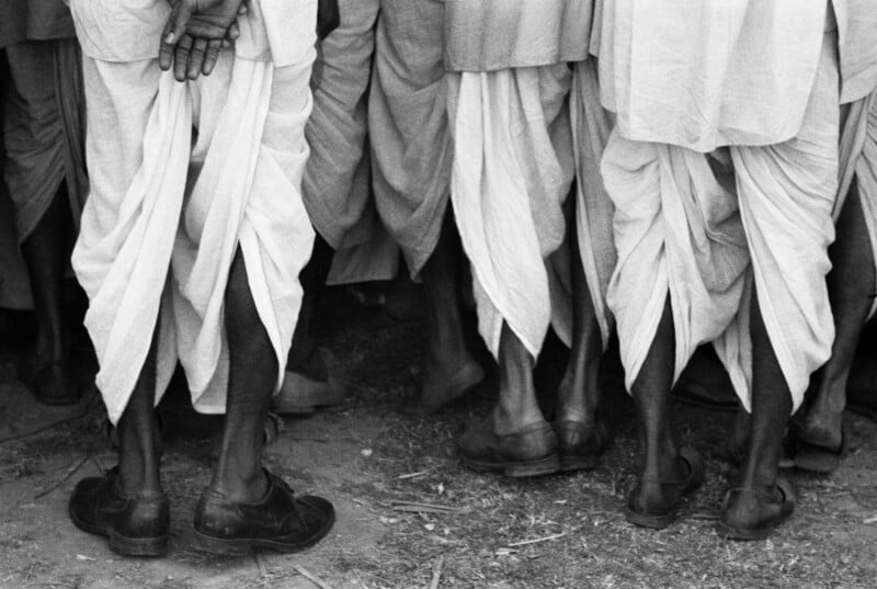 A low-angle, black-and-white shot showing the lower bodies and legs of several men wearing traditional dhotis and dark leather shoes.