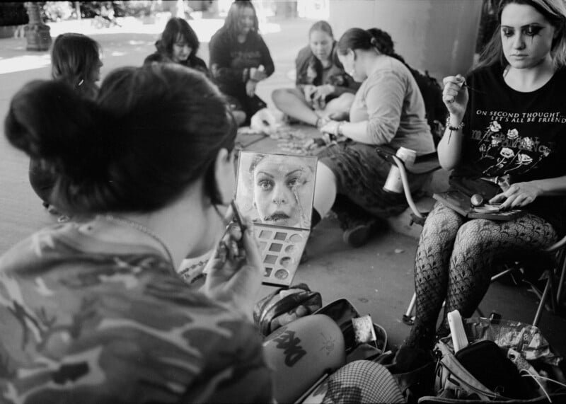 A group of young women sit together outdoors, applying makeup and preparing for an event.
