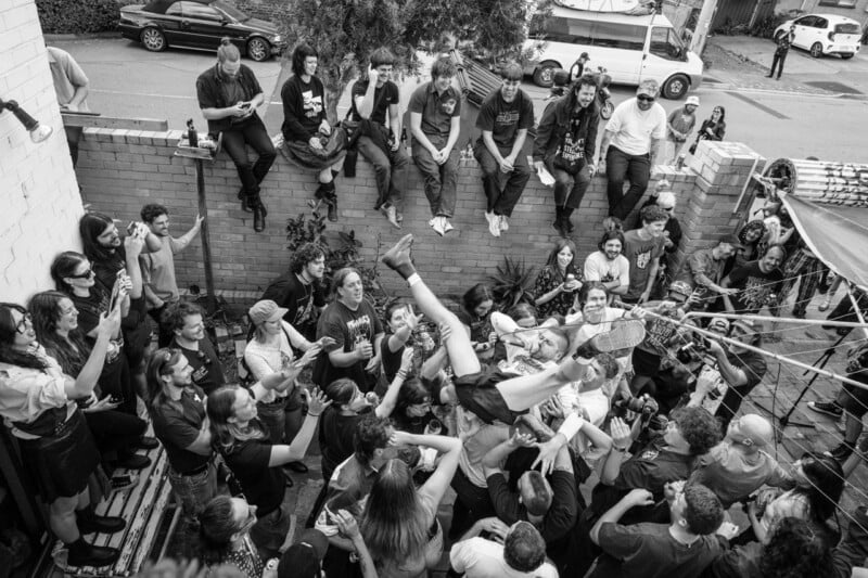 A crowd of people gathers in an outdoor space, with several individuals sitting on a brick wall while others hold a person aloft in the center.