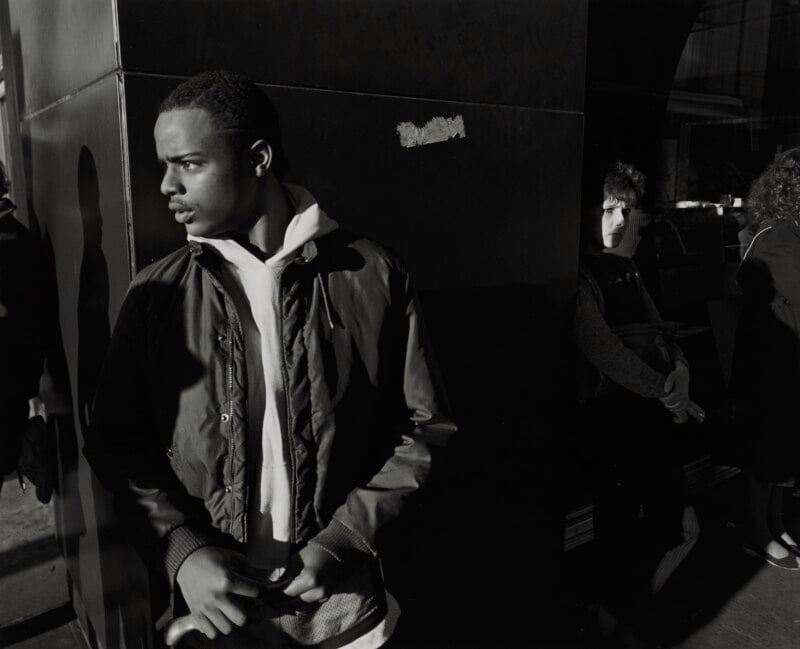 A young man in a jacket and hoodie stands in the foreground, looking to the left, while a person stands in the shadows behind him.