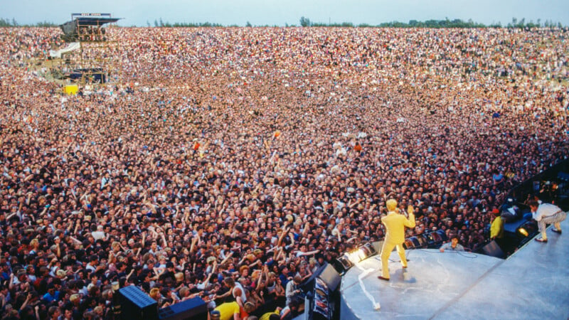 A performer in a yellow suit stands on a stage facing a massive crowd of people stretching across a large outdoor field.