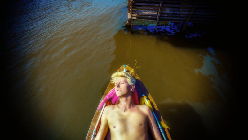 A shirtless person with blonde hair lies with eyes closed in a small wooden boat on murky water near a wooden structure.