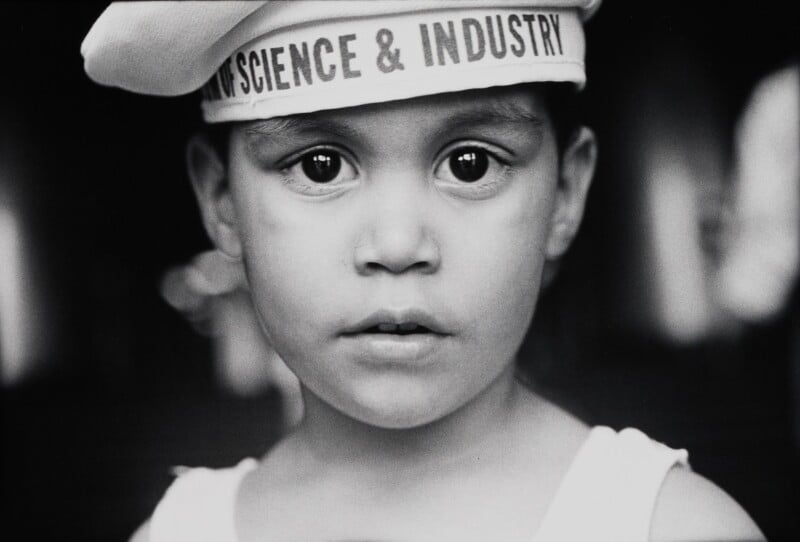 A close-up, black-and-white portrait of a young child wearing a white sailor-style cap labeled "SCIENCE & INDUSTRY.