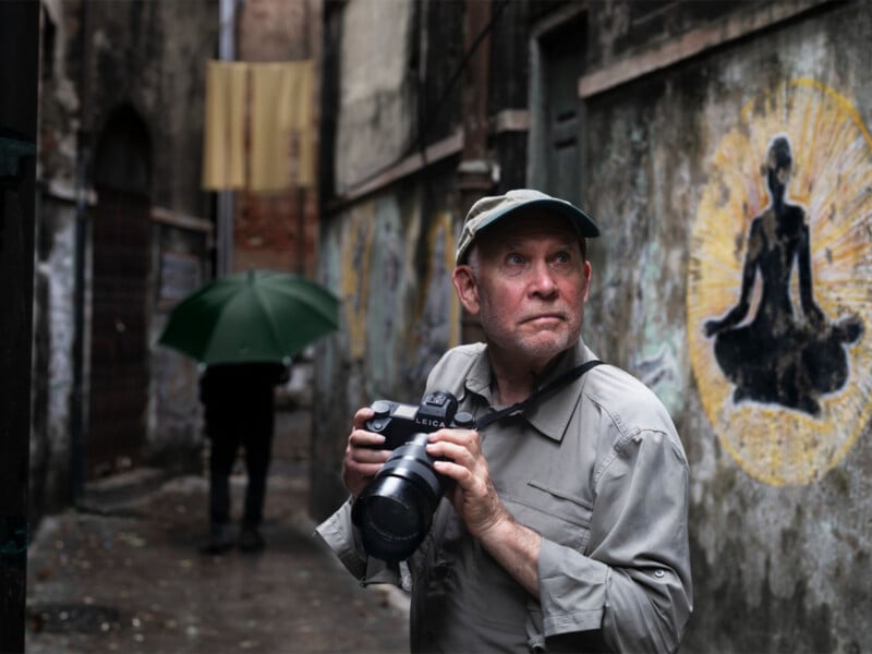 A man in a cap and light-colored shirt holds a camera while standing in a narrow alleyway with a mural of a seated figure.