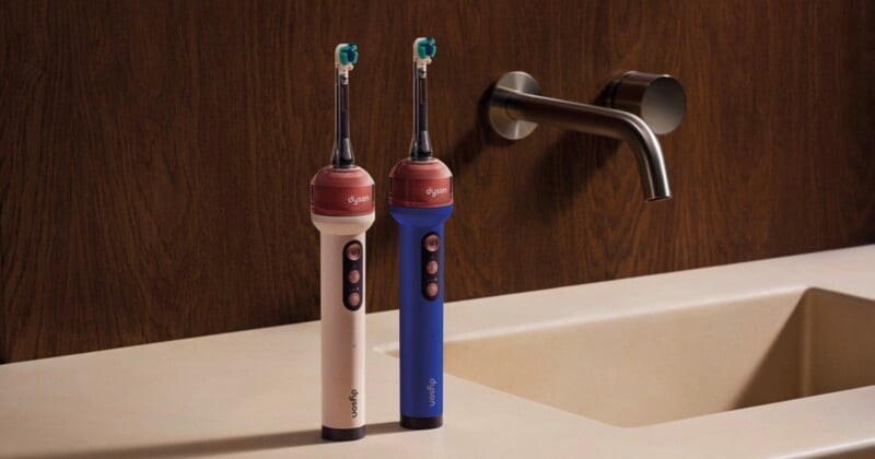 Two Dyson electric toothbrushes, one cream and one blue, stand on a bathroom counter next to a sink faucet.