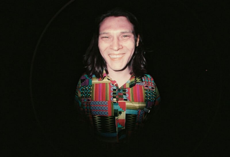 A person with long dark hair and a wide smile wears a colorful, patterned button-down shirt against a dark background.