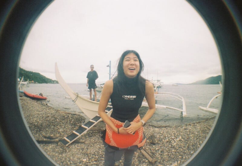 A woman in a black sleeveless wetsuit stands on a rocky beach, with a man standing on a boat behind her.
