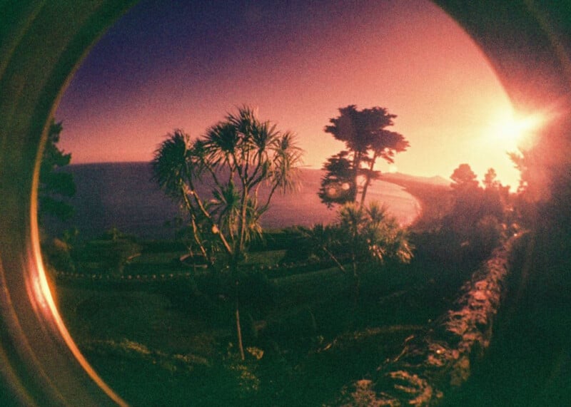 A circular, vignette-framed view of a coastal landscape at sunset, featuring palm-like trees, a dark shoreline, and a bright sun on the horizon.