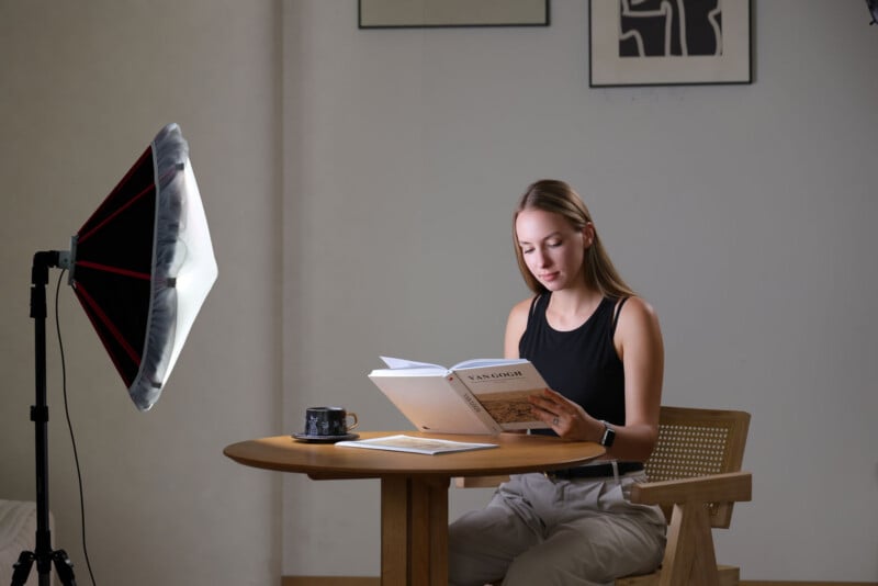 A woman sits at a round wooden table reading a Van Gogh book, illuminated by a large studio softbox light on a stand.
