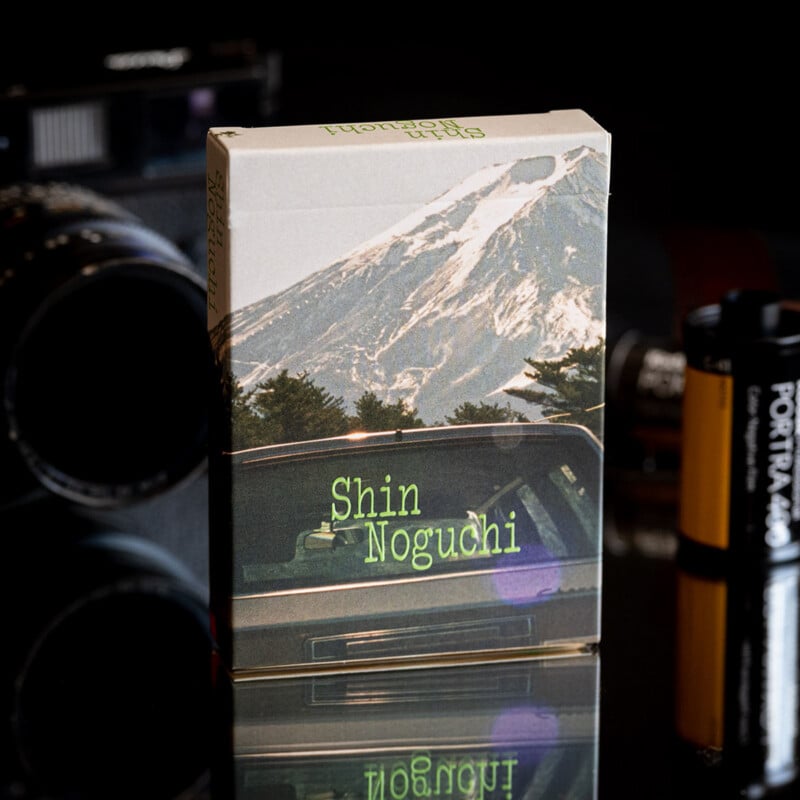 A deck of playing cards featuring a mountain landscape and a car on the box sits on a reflective surface next to a camera lens and a roll of film.