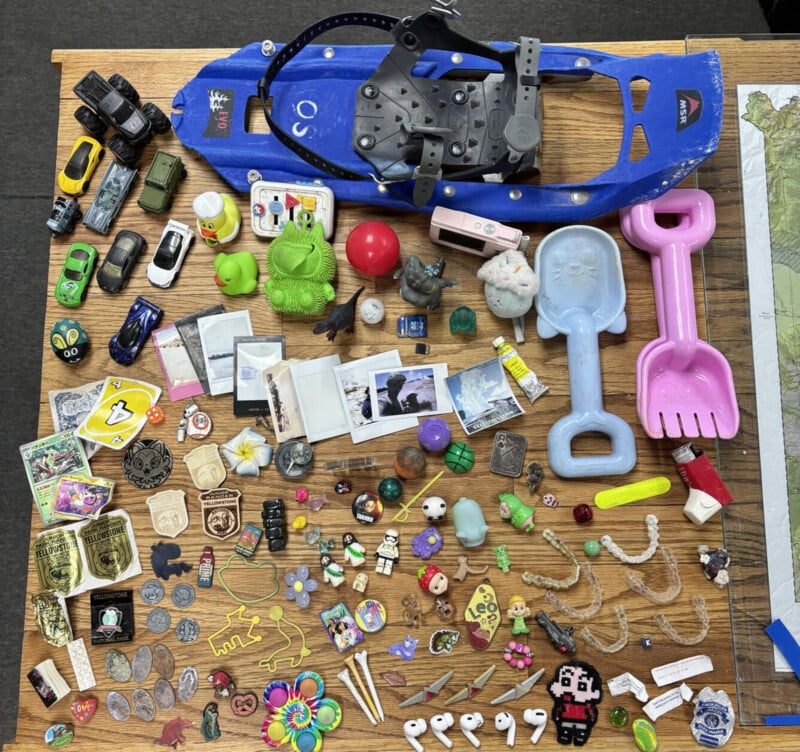 A collection of miscellaneous items including a blue snowshoe, toy cars, small figurines, instant photos, pins, coins, and plastic dental aligners spread across a wooden table.