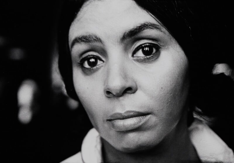 A close-up, black-and-white portrait of a woman with dark hair looking directly at the camera with a neutral expression.