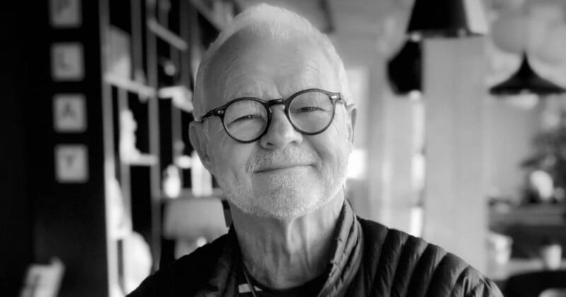 A black-and-white portrait of an older man with white hair and a beard, wearing round glasses and a quilted jacket.
