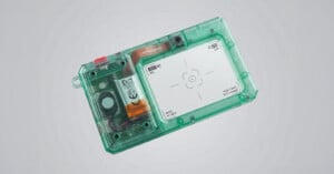 A translucent green rectangular device with a visible internal battery, circuit board, and a white screen featuring a centered crosshair target.