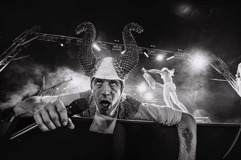 A performer wearing a spiked horned headpiece and costume leans over a stage barrier, holding a microphone and looking upward.