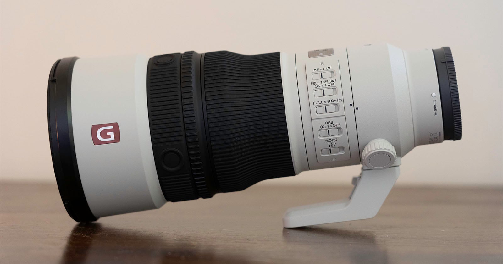 A white telephoto camera lens with a black zoom ring and a series of control switches on the side, resting on a wooden surface.