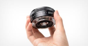 A hand holds a black camera lens, showing the rear mount, electronic contacts, and an aperture ring marked with f-stop numbers.