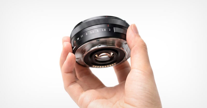 A hand holds a black camera lens, showing the rear mount, electronic contacts, and an aperture ring marked with f-stop numbers.