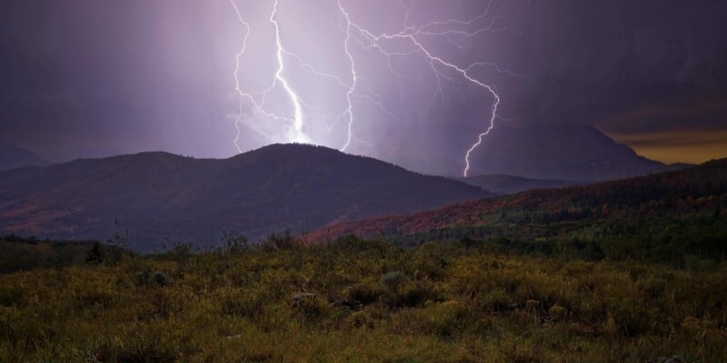 Two bright lightning bolts strike the peaks of dark, forested mountains at night above a grassy foreground.