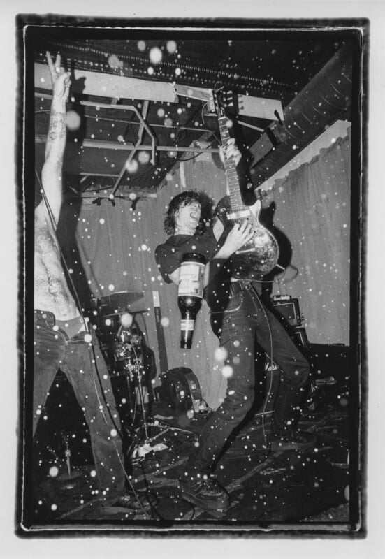 A black-and-white concert scene shows a guitarist playing while holding a beer bottle, with another person standing beside them with an arm raised.