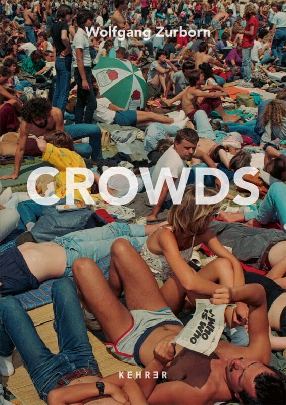 A dense crowd of people sits and lies on a grassy field, with a person in the foreground reading a newspaper titled "WHO IS WHO.