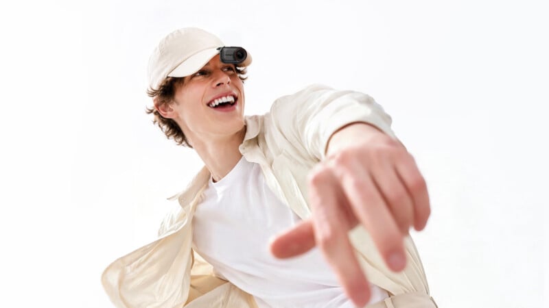 A smiling young man wearing a light-colored cap with a small camera attached to the brim reaches his hand toward the camera against a white background.