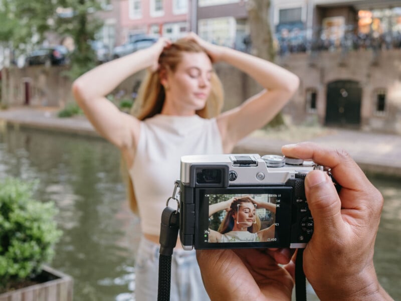 A person holds a digital camera to photograph a woman posing with her hands in her hair by a canal.
