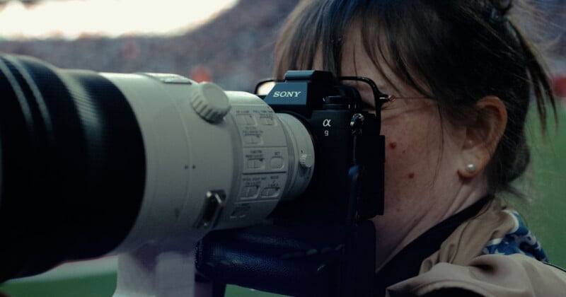 A person with dark hair and glasses looks through the viewfinder of a Sony Alpha 9 camera with a large telephoto lens.