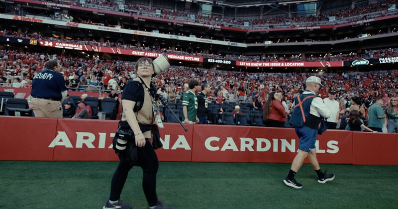 A photographer carrying a large camera lens walks along the sideline of a crowded football stadium in front of a red Arizona Cardinals wall.