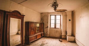 An abandoned room with peeling white walls, a wooden wardrobe with a mirror, a glass-fronted cabinet, and a hanging lamp.