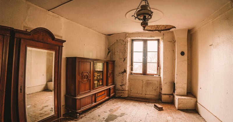 An abandoned room with peeling white walls, a wooden wardrobe with a mirror, a glass-fronted cabinet, and a hanging lamp.
