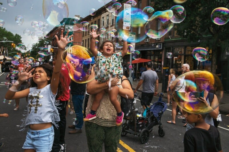 A young girl is held up by an adult while another girl reaches toward many large, iridescent bubbles floating in a city street.