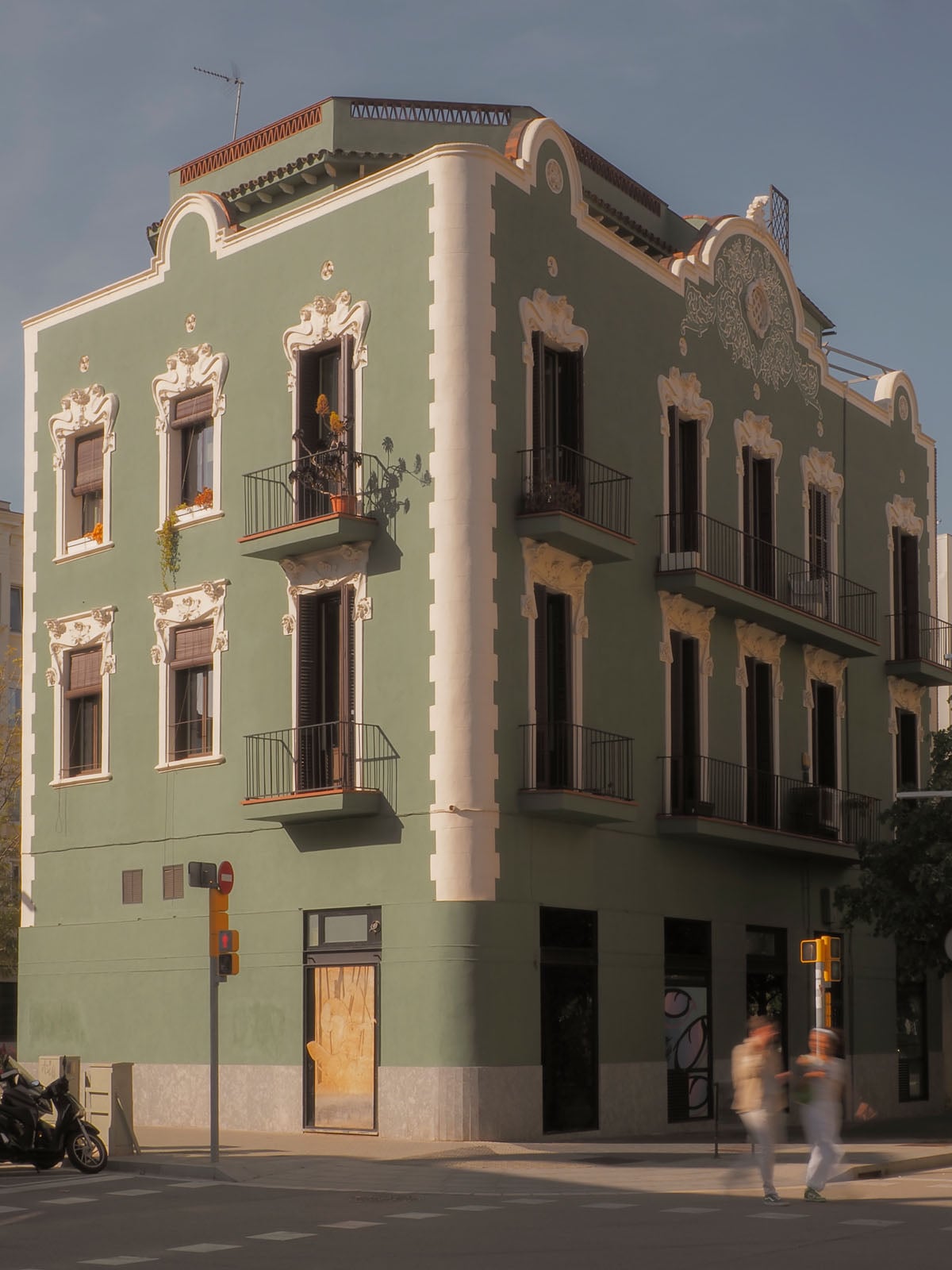 A three-story green corner building with white trim, ornate window moldings, and black iron balconies, situated at a street intersection with two blurred pedestrians walking by.