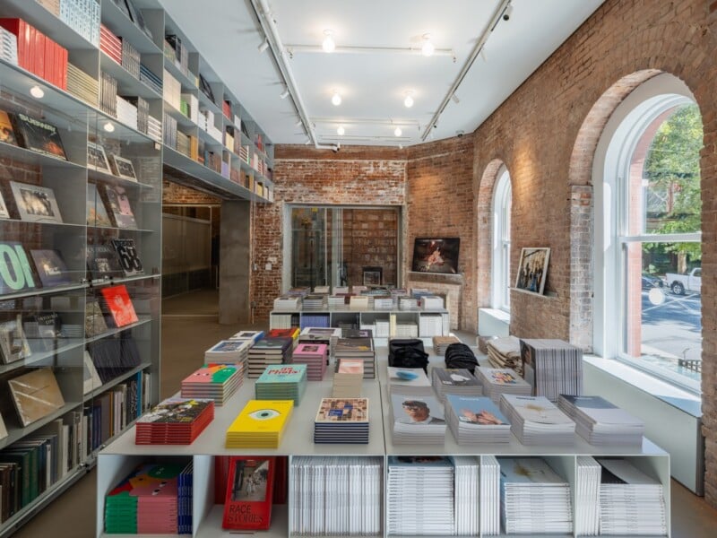A bright bookstore interior features floor-to-ceiling shelves filled with books, a central display table stacked with magazines and art books, and arched windows set into exposed brick walls.