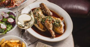 A roasted half chicken topped with chimichurri in a brown sauce, served alongside a side salad, fries, and a glass of white wine on a wooden table.