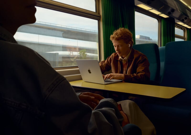 A person with curly red hair sits at a train table working on a laptop while wearing wireless earbuds.
