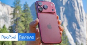 A hand holds a dark red smartphone with a prominent, raised camera module against a blurred mountain landscape background.
