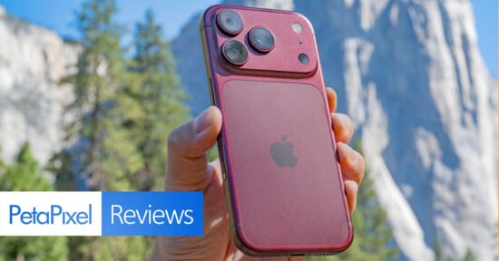 A hand holds a dark red smartphone with a prominent, raised camera module against a blurred mountain landscape background.