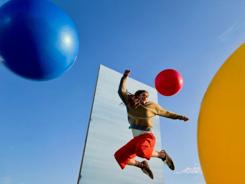 A person with long hair jumps in the air in front of a tall, rectangular mirror, surrounded by large blue, red, and yellow spheres against a clear blue sky.