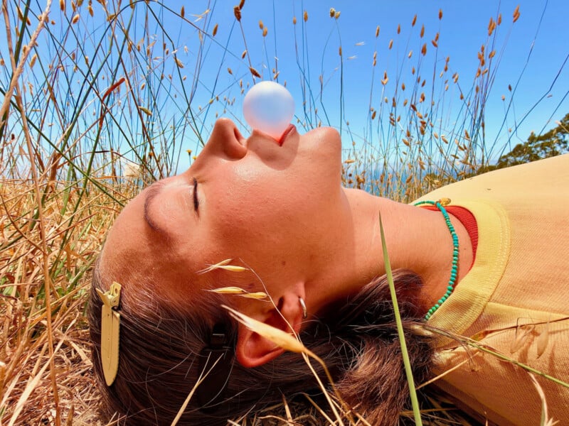 A person with closed eyes lies in tall, dry grass under a clear blue sky, blowing a bubble from their mouth.