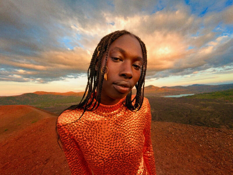 A person with braided hair and gold hair accessories wears a shimmering orange sequined top while standing on a rocky, reddish landscape at sunset.